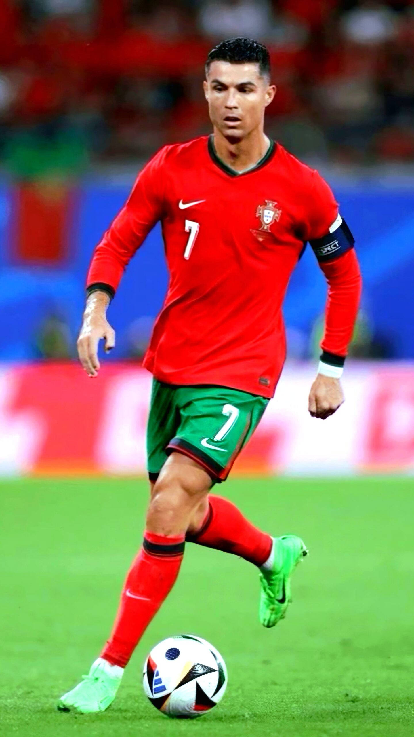 A male soccer player in a red and green Portugal uniform, wearing number 7 and a captain's armband, is running on the field with the ball at his feet. Blurred spectators and a blue banner hint at live coverage, perfect for enjoying on IPTV French channels.
