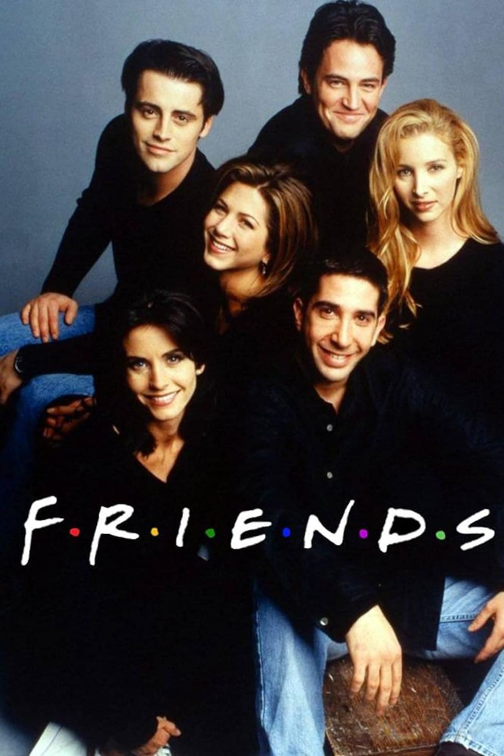 Six people sit and smile together in a group, wearing casual dark-colored clothes, in front of a gray background. The word FRIENDS appears at the bottom in white playful letters with colorful dots between each letter, capturing the spirit of connection—much like enjoying your favorite shows on IPTV FRENCH.